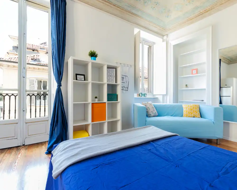 Camera da letto luminosa e moderna con copriletto blu, divano azzurro e grandi finestre che garantiscono molta luce naturale. Scaffalatura decorativa e soffitto dipinto aggiungono carattere, creando uno spazio accogliente.