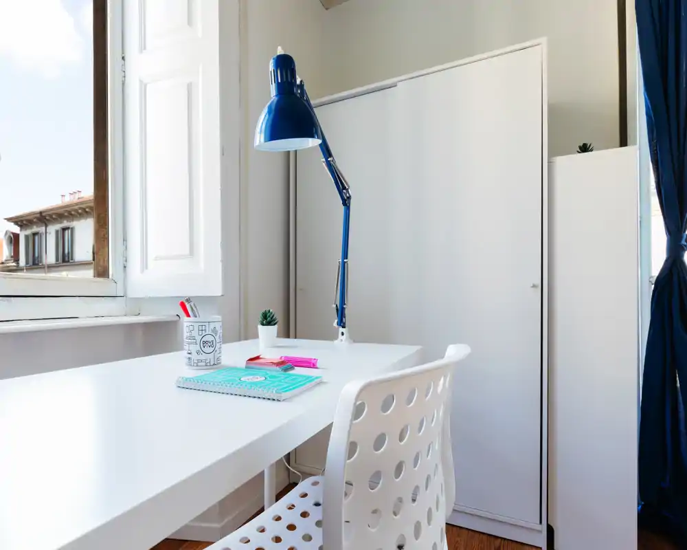 Angolo studio luminoso e ordinato con scrivania bianca, sedia traforata e lampada da tavolo blu vicino a una finestra aperta — ideale per lavoro o lettura.