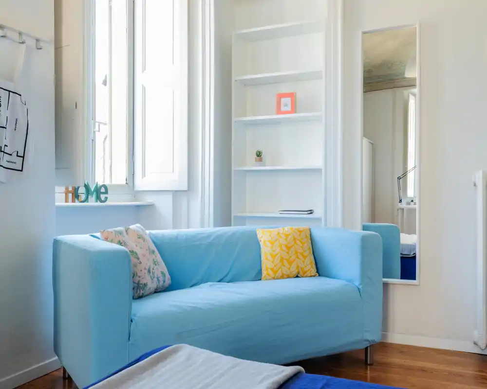 Zona giorno luminosa e moderna con divano azzurro, cuscini decorativi e luce naturale dalla finestra — un angolo accogliente per rilassarsi.