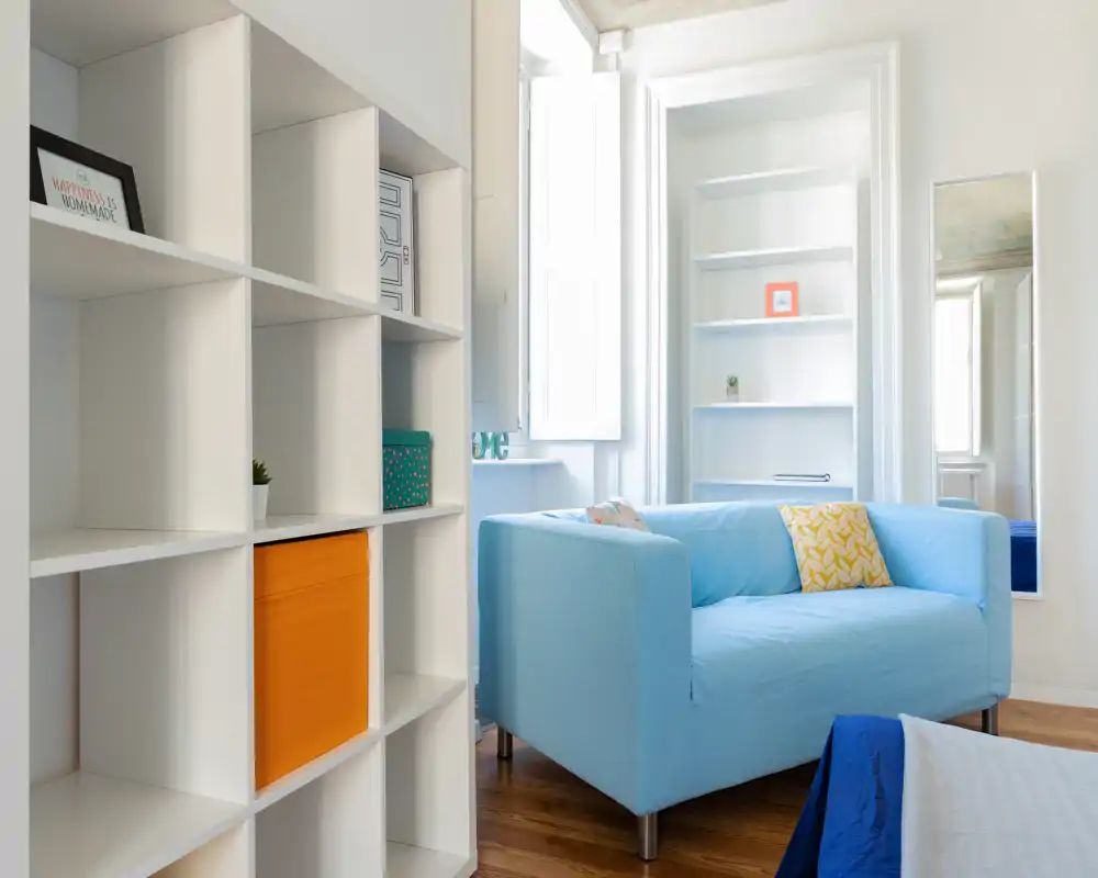 Zona giorno luminosa e moderna con divano azzurro, scaffalatura e luce naturale da una finestra alta — perfetta per un monolocale accogliente.