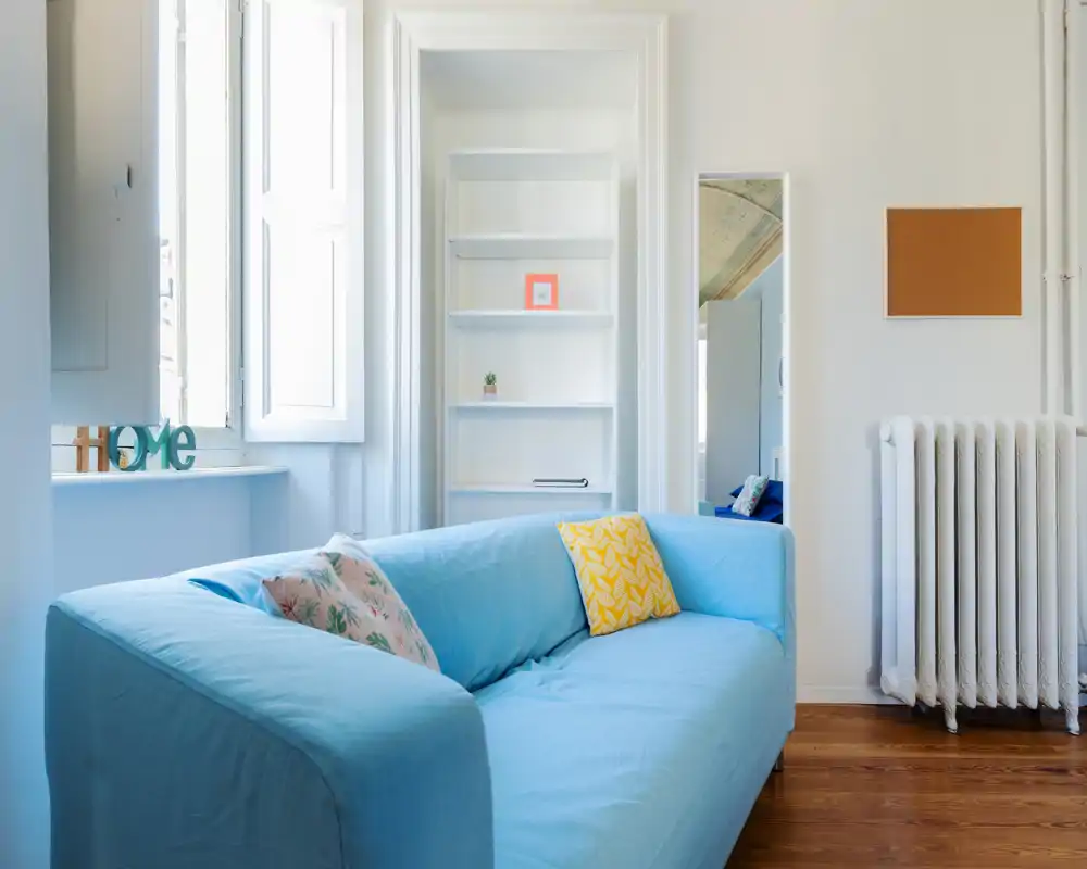 Soggiorno luminoso e arioso con divano azzurro, cuscini decorativi e luce naturale dalle grandi finestre — stile pulito e minimale, perfetto per affitti.