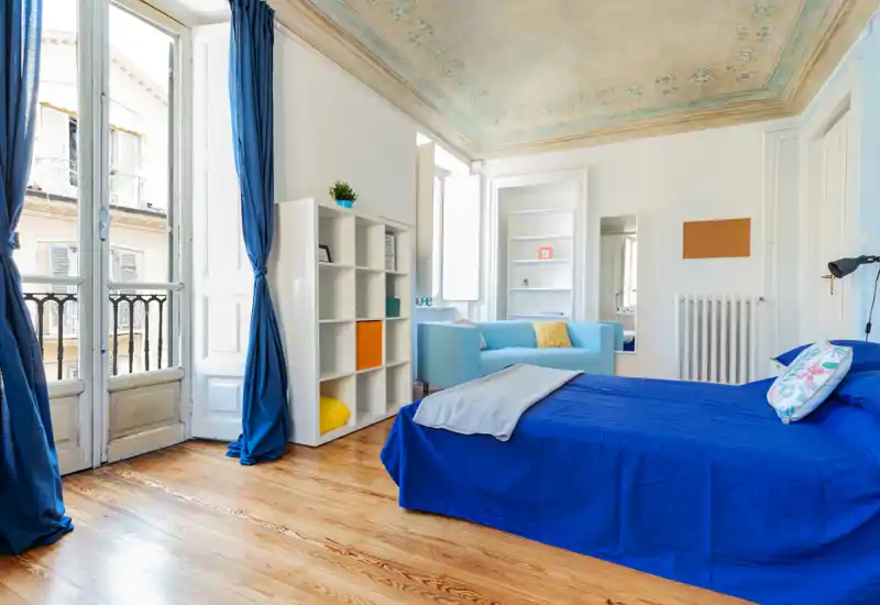 Camera da letto luminosa con letto in blu, tende coordinate e un piccolo divano; soffitti alti con decorazioni a fresco e pavimenti in legno conferiscono un'atmosfera ariosa ed elegante.