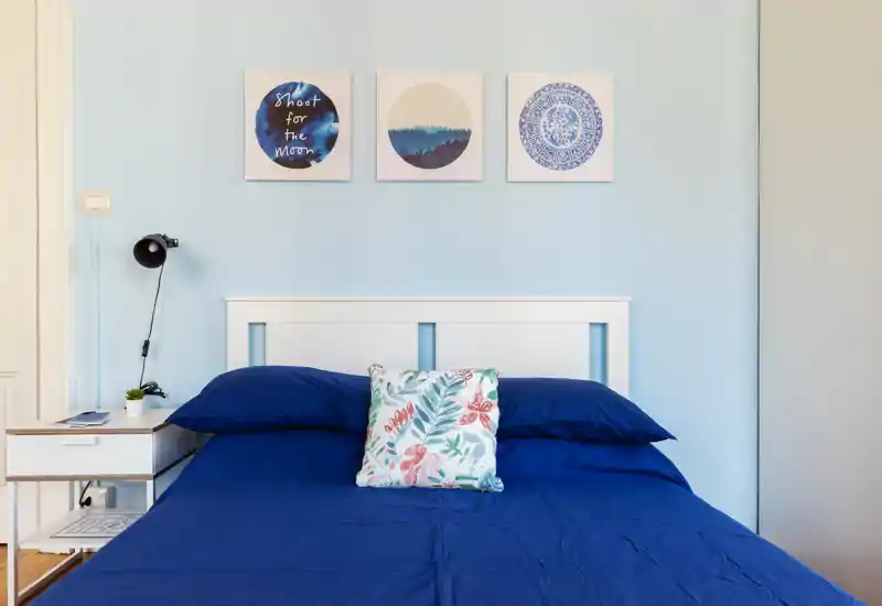 Camera da letto luminosa e ordinata con copriletto blu navy, cuscino decorativo floreale e quadri minimal sopra la testata. Un piccolo comodino con lampada e piantina completa l'atmosfera calma e accogliente, ideale per annunci.