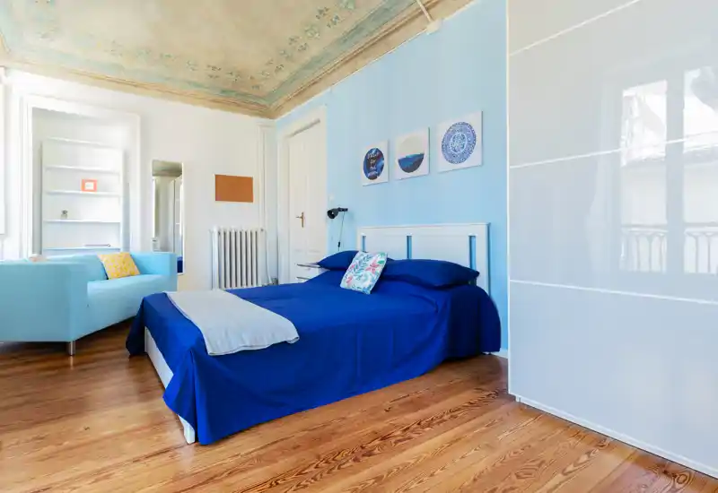 Camera da letto luminosa e di stile con letto matrimoniale blu, cuscini decorativi e un'area salotto; pavimento in legno e soffitto decorato aggiungono carattere.
