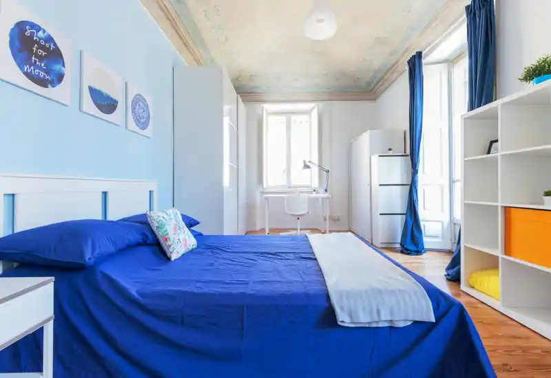 Camera da letto luminosa e ariosa con copriletto blu intenso, cuscini decorativi e scrivania vicino alla finestra. Soffitti alti con decorazioni e pavimenti in legno caldo rendono l'ambiente accogliente.