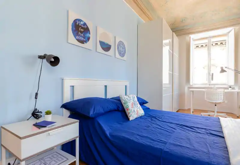 Camera da letto luminosa e moderna con letto matrimoniale con biancheria blu, comodino e una postazione di lavoro vicino alla finestra. Luce naturale e quadri alle pareti rendono l'ambiente accogliente.