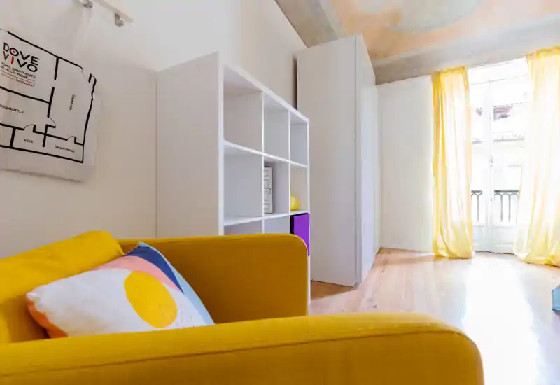 Zona giorno moderna e luminosa con un divano giallo vivace, cuscini decorativi e una libreria a cubi. Ampia finestra/porta lascia entrare molta luce naturale mettendo in risalto il parquet e il soffitto affrescato.
