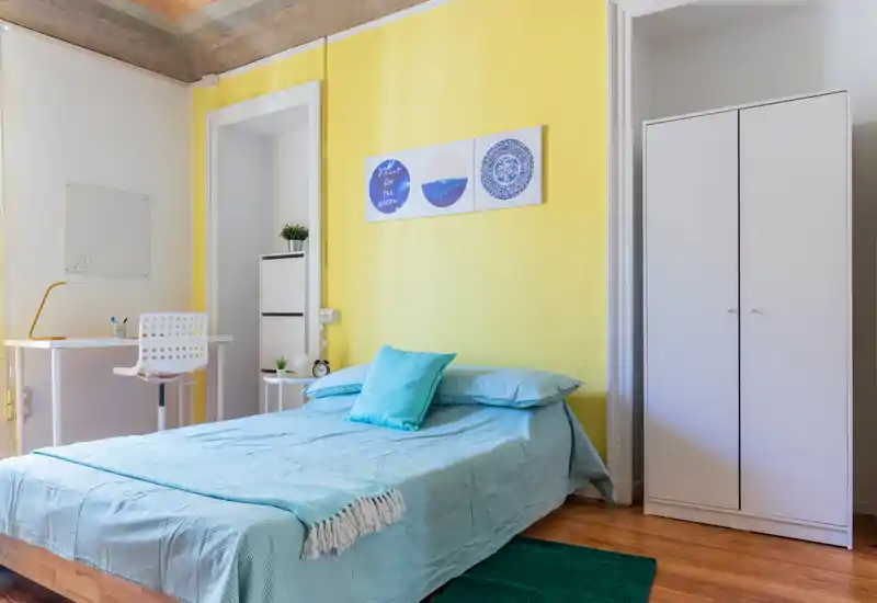 Camera da letto luminosa e curata con letto matrimoniale con biancheria turchese, angolo studio e armadio bianco su una parete accentata gialla.