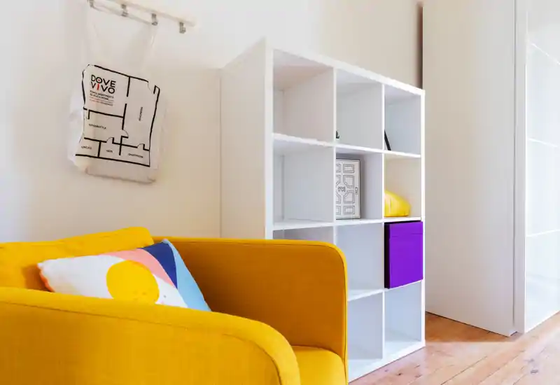 Zona giorno moderna e luminosa con poltrona giallo senape e scaffalatura bianca modulare; cuscino colorato e allestimento curato rendono l'angolo accogliente.