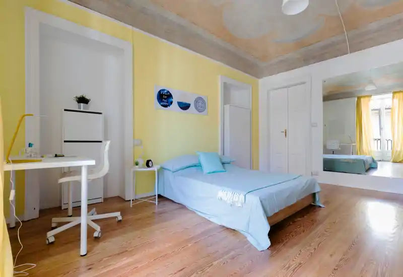 Camera da letto luminosa e ariosa con letto matrimoniale in lenzuola azzurre, piccolo comodino, scrivania e grande specchio a parete che riflette la luce naturale della finestra.