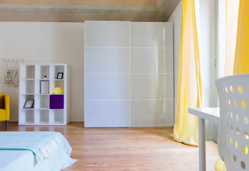 Camera da letto moderna e luminosa con angolo del letto visibile, armadio scorrevole lucido, pavimenti in legno e tende gialle che danno calore.