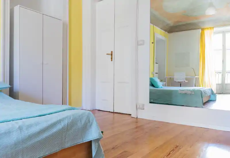 Camera da letto luminosa e ariosa con letto basso con biancheria turchese, pavimento in legno chiaro e grande specchio che riflette la stanza. Accenti gialli e ampia finestra donano luce naturale calda, ambiente minimal e accogliente.