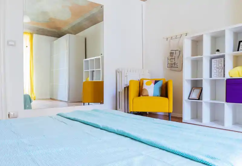 Camera da letto luminosa e moderna con copriletto azzurro, poltrona gialla come accento e scaffalatura ordinata; luce naturale e colori vivaci creano un ambiente accogliente.