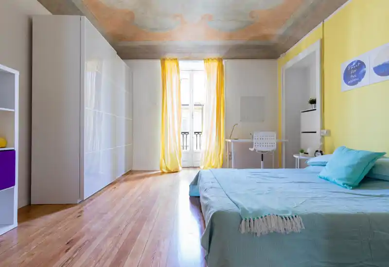 Camera da letto luminosa e raffinata con letto matrimoniale con biancheria azzurra, una scrivania vicino alla finestra e ampi armadi bianchi. Pavimento in legno, tende gialle e un soffitto decorato che dona carattere.