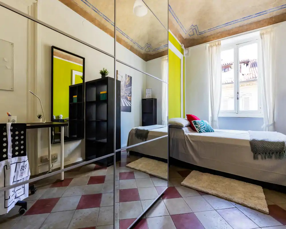 Camera singola luminosa e ben arredata con letto vicino a una grande finestra, cuscini decorativi e morbidi tappeti; gli armadi specchiati ampliano lo spazio.