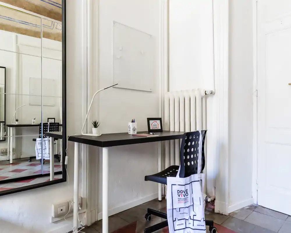 Zona lavoro luminosa e minimal in un soggiorno con scrivania semplice, sedia, lampada e un grande specchio che dona profondità e luce.
