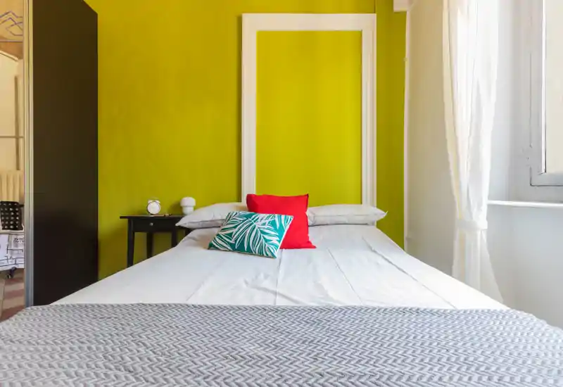 Chambre lumineuse et élégante avec un lit double centré, coussins colorés et lumière naturelle par la fenêtre.