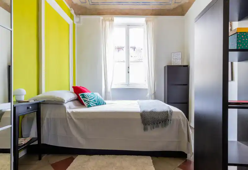 Camera da letto luminosa e ben allestita con un letto singolo, cuscini colorati e luce naturale dalla finestra centrale. Dettagli curati come il plaid, il comodino e il tappeto rendono l'ambiente accogliente.