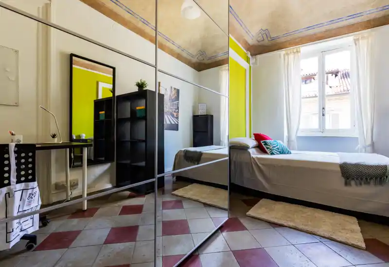 Camera singola luminosa e di stile con letto ordinato vicino a una grande finestra, armadio con ante a specchio e soffitto decorativo—perfetta per un soggiorno accogliente in città.