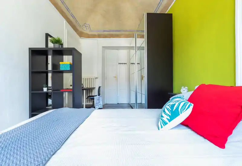 Camera da letto luminosa e alla moda con un ampio letto e cuscini colorati, un armadio con ante specchiate e scaffalatura aperta per riporre gli oggetti. Soffitti alti e dettagli decorativi rendono la stanza accogliente.