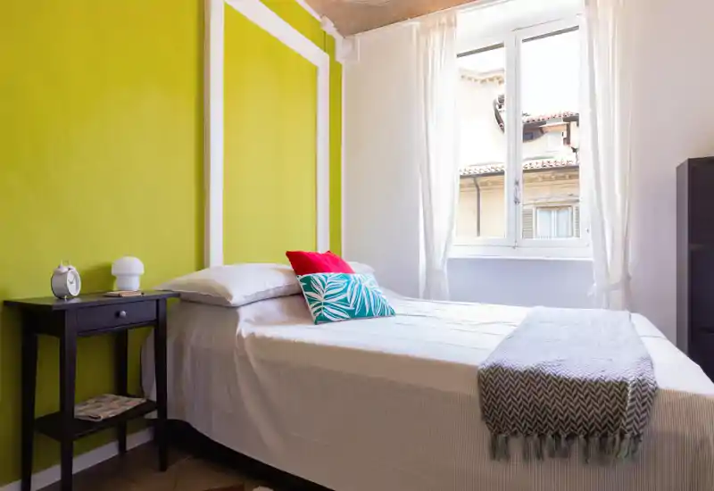 Camera da letto singola luminosa e ben arredata con parete accent verde, biancheria bianca, cuscini decorativi e comodino. La luce naturale dalla finestra rende l’ambiente arioso e accogliente, perfetto per annunci.