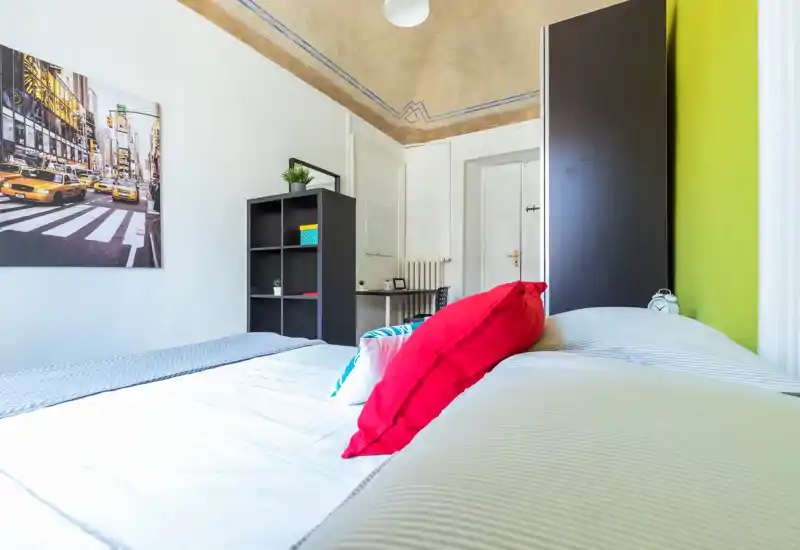Camera da letto luminosa e ben arredata con un ampio letto in primo piano, cuscini colorati e scaffalatura e scrivania semplici sullo sfondo.