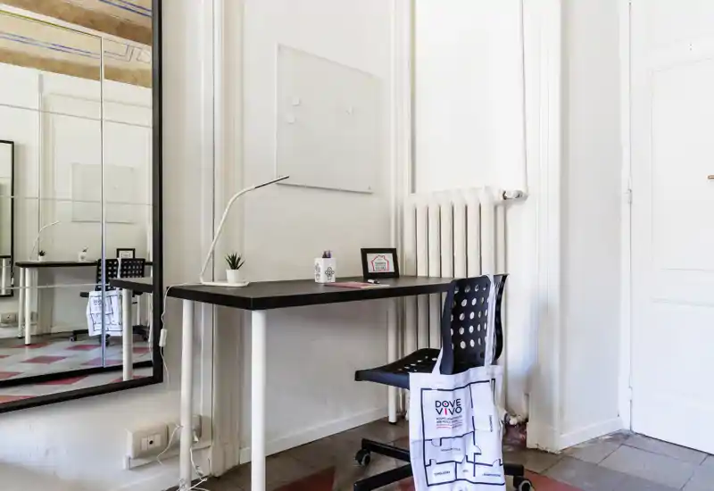 Zona lavoro minimal e luminosa con una scrivania nera compatta, lampada da tavolo moderna e sedia forata davanti a pareti bianche e un grande specchio che riflette l'ambiente.