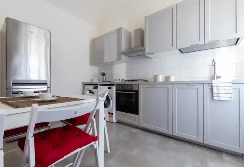 Cucina luminosa e moderna con mobili grigio chiaro, forno incassato e piano cottura a gas, frigorifero in acciaio e tavolo da pranzo con sedie con cuscini rossi — perfetta per annunci di appartamenti compatti.