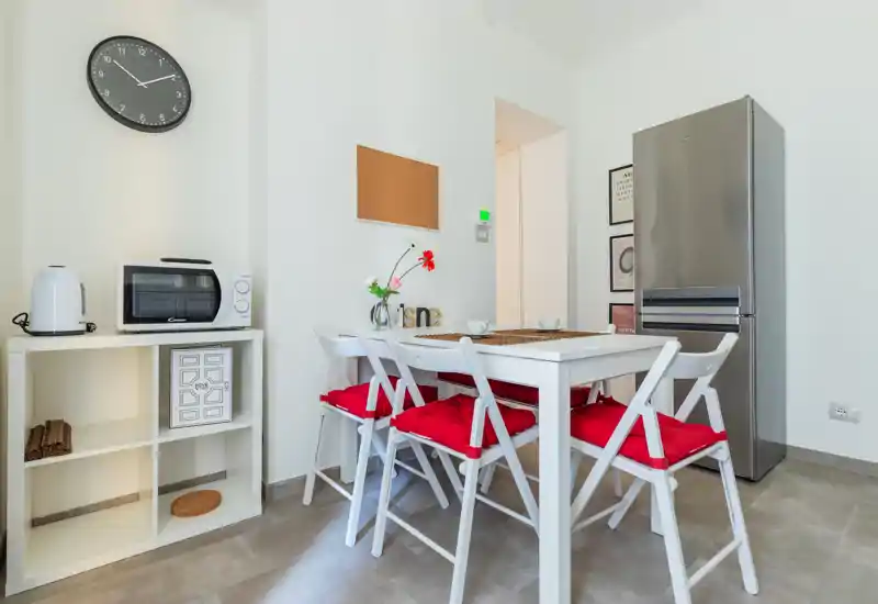 Zona pranzo moderna e luminosa con tavolo bianco, quattro sedie con cuscini rossi e frigorifero in acciaio inox; tocchi decorativi semplici come un vaso con fiori e stampe incorniciate.