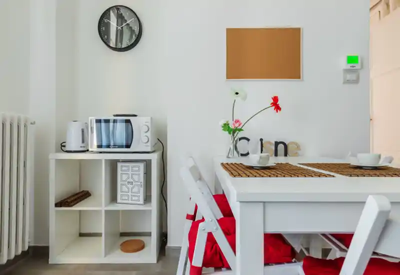 Angolo pranzo luminoso e moderno con tavolo bianco, cuscini rossi, tovagliette e fiori decorativi — ideale per pasti informali.