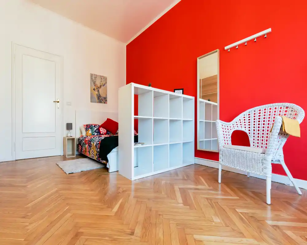 Chambre lumineuse et moderne avec lit simple coloré, meuble étagère blanc en séparation et fauteuil en osier devant un mur rouge vif — un espace compact et stylé, idéal pour les locations courtes.