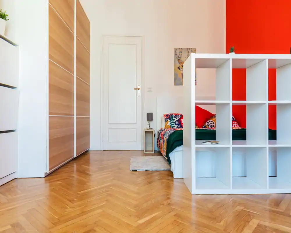 Coin chambre lumineux et moderne avec un lit coloré et une étagère blanche servant de séparateur. Parquet en chevron, grand placard et lampe de chevet minimal complètent l'espace aérien.