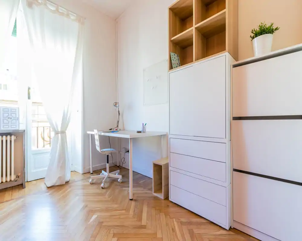 Espace de vie lumineux et minimaliste avec bureau blanc, chaise pivotante et rangements hauts; grande porte-fenêtre apportant de la lumière naturelle et parquet chaleureux.