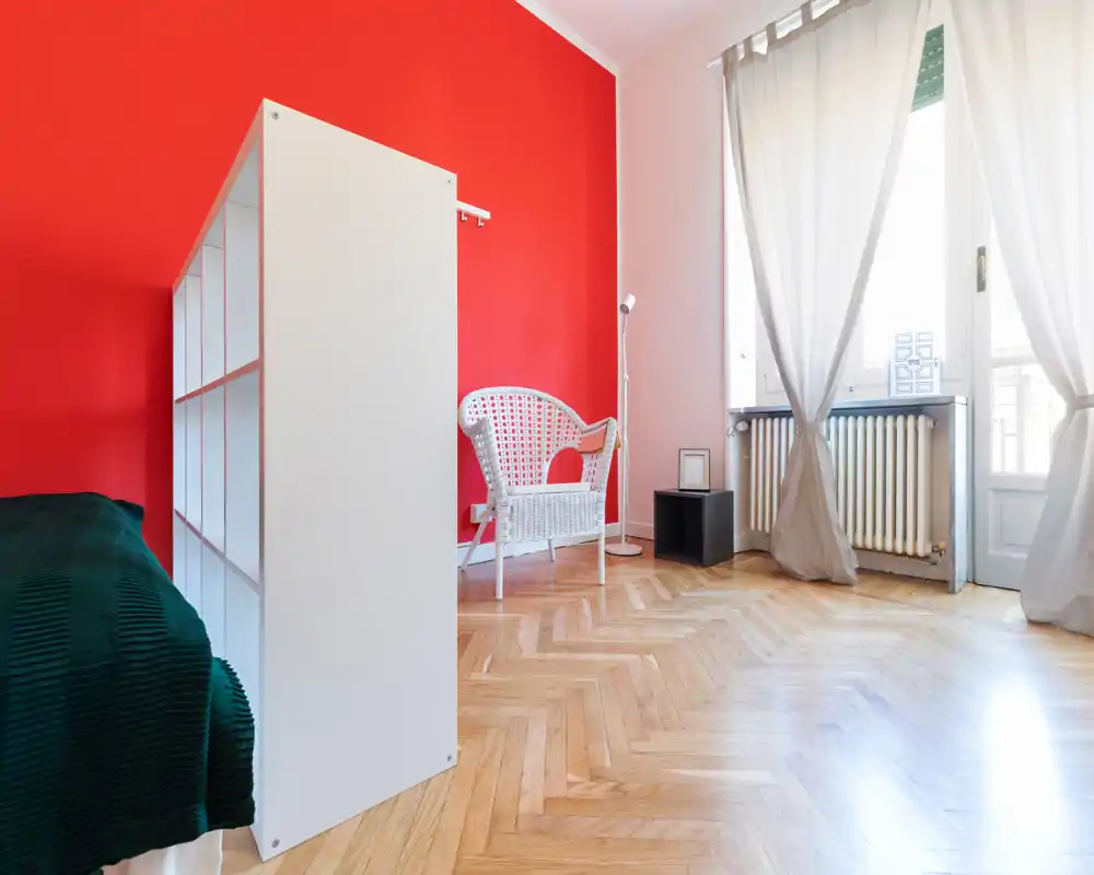 Chambre lumineuse avec un mur rouge vif, parquet et lumière naturelle provenant d'une grande fenêtre. Le lit est partiellement visible à gauche ; une chaise en osier blanche et des étagères minimales créent une atmosphère aérée.
