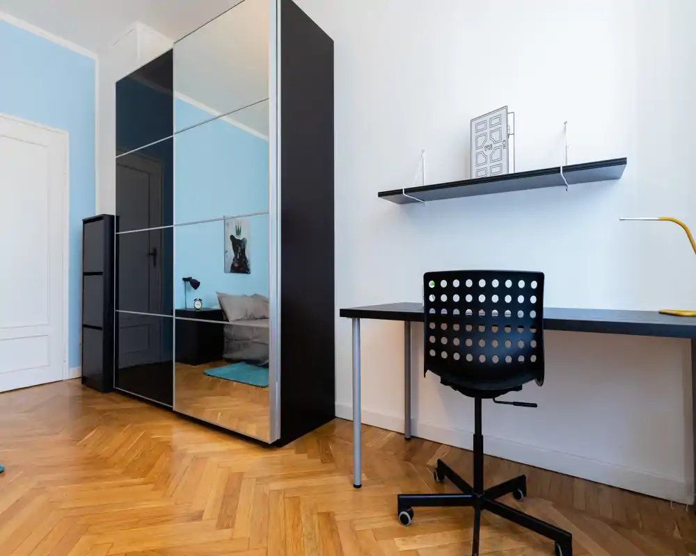 Dormitorio moderno y luminoso con armario con espejo, zona de cama visible y un escritorio minimalista. Suelo de parquet y detalles acogedores que hacen la estancia funcional para alquileres.