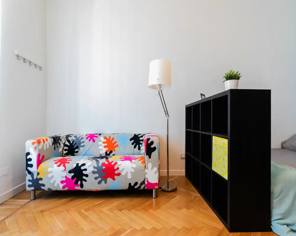 Zona de estar moderna y luminosa con un sofá estampado colorido, lámpara de pie y estantería que separa el espacio — ideal para un estudio con estilo.