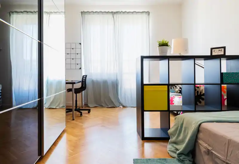 Dormitorio moderno y luminoso con cama bien hecha, estantería divisoria y amplias ventanas que aportan luz natural.