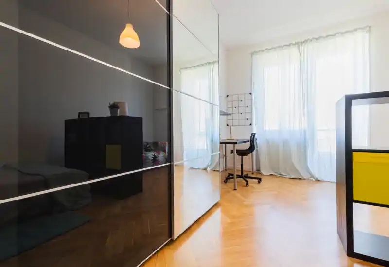 Dormitorio moderno y luminoso con grandes ventanas, cortinas translúcidas y suelo de parquet. Un pequeño escritorio y silla junto a la ventana; la cama se ve parcialmente a la izquierda junto a un mueble, dando una sensación limpia y aireada.