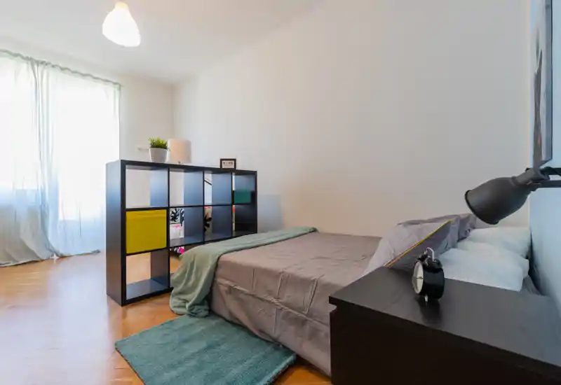 Dormitorio moderno y luminoso con cama baja de matrimonio, mesita, lámpara y estantería abierta como separador. La luz natural y los textiles suaves crean un ambiente acogedor.