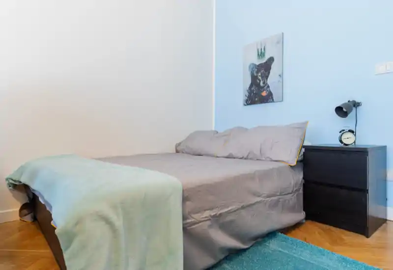 Dormitorio ordenado y compacto con cama doble vestida en gris, mesita con lámpara y reloj, toques turquesa y suelo de madera. Luminoso, moderno y acogedor, perfecto para alquileres de corta estancia.