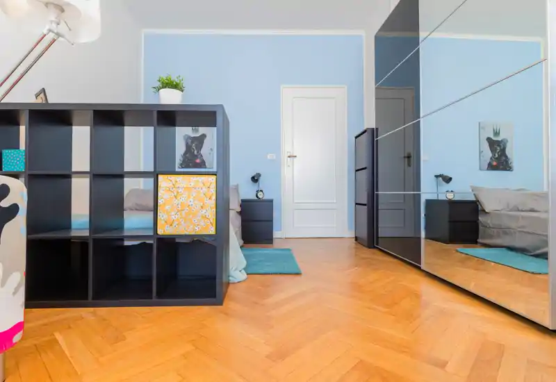 Dormitorio moderno y luminoso con pared azul, armario con puertas de espejo y cama baja parcialmente visible junto a una mesita negra y una alfombra turquesa — limpio y con estilo.