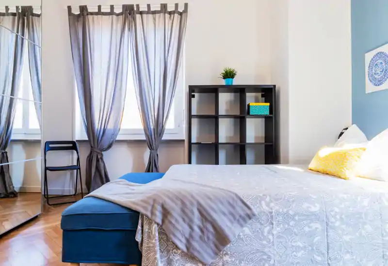 Bright, well-styled bedroom with a large bed dressed in patterned linens, a blue bench at the foot, and tall windows with sheer curtains that let in soft natural light.