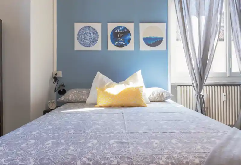 Dormitorio luminoso y bien decorado con cama grande centrada frente a una pared azul, cojines decorativos y cuadros; luz natural abundante por la ventana con cortinas claras.