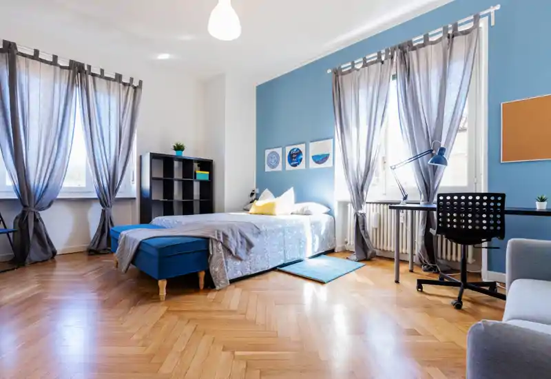 Dormitorio luminoso y con estilo, pared azul, grandes ventanas con cortinas semitransparentes, cama doble cómoda con cojines y un escritorio junto a la ventana — ideal para un alquiler moderno.