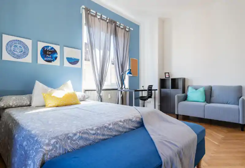 Dormitorio luminoso y aireado con cama completa, pared azul de acento y pequeño espacio con sofá y escritorio. La luz natural entra por la ventana; los textiles y cuadros ofrecen un ambiente acogedor, perfecto para un anuncio.