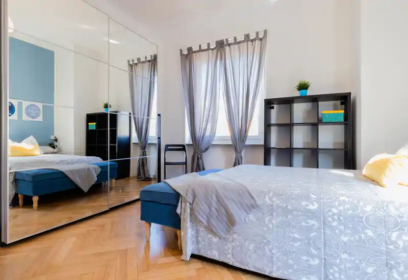 Bright, modern bedroom with a full bed, mirrored wardrobe and parquet flooring; airy curtains and calm blue accents create a relaxing atmosphere.