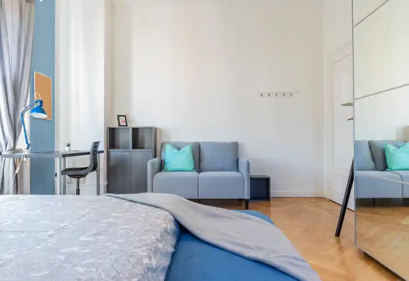 Bright, modern bedroom with a comfortable bed in the foreground, a small sofa and desk by the window; neutral tones and teal accents create a calm, airy atmosphere.