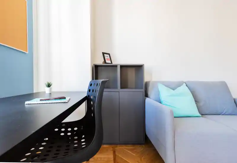 Bright, modern living area with a compact sofa, decorative cushion and a workspace desk—clean lines and soft natural light create a comfortable, functional corner ideal for short stays.
