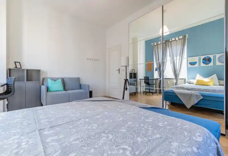 Dormitorio moderno y luminoso con cama grande, armario con espejo y escritorio junto a la ventana. Tonos neutros con acentos azules y cojines que crean un ambiente acogedor.