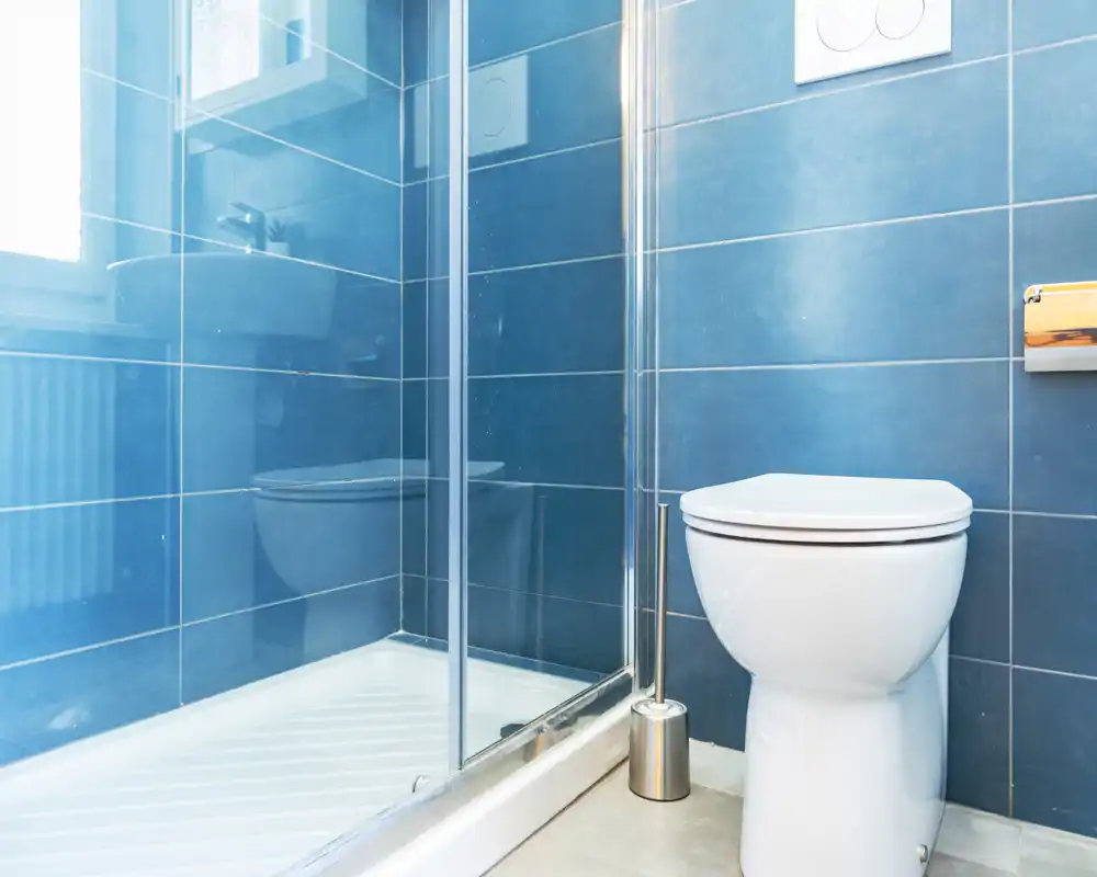 Baño moderno y luminoso con paredes azules alicatadas, cabina de ducha de cristal y inodoro blanco; líneas limpias y suelos neutros para un espacio funcional.